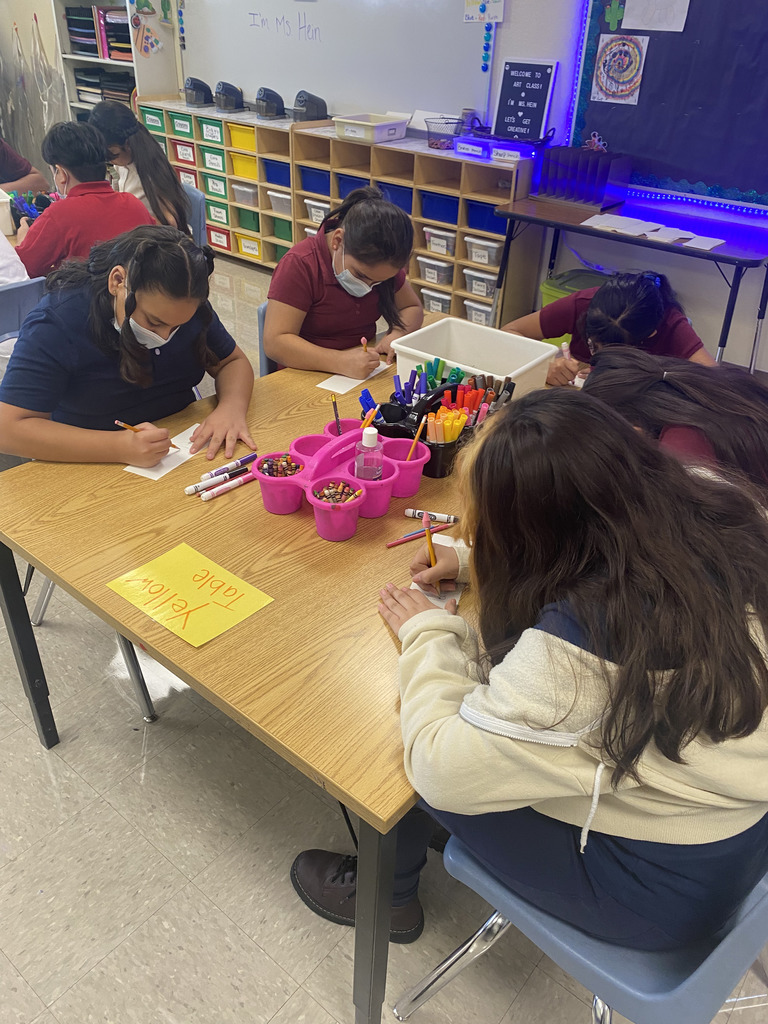 5th grade students in art class sitting at a table drawing on a piece of paper and colored pencils