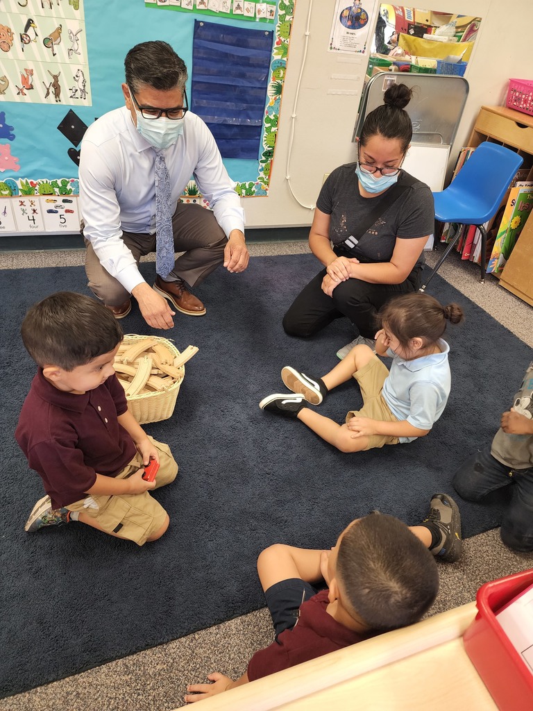 preschool students sitting on a carpet with the teacher and superintendent practicing how to sit down