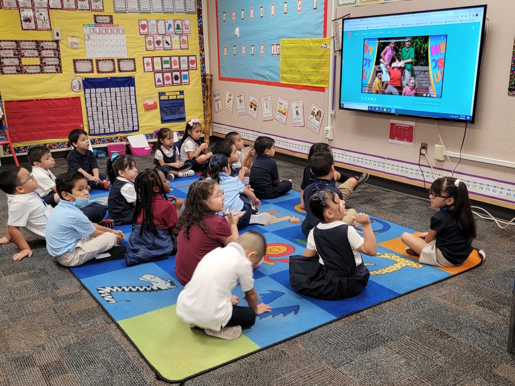 elementary students sitting on the classroom carpet looking at the screen to follow along with the teacher's lesson