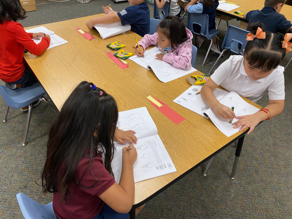 5 kindergarten students focused on their worksheet by tracing the shapes on their page.
