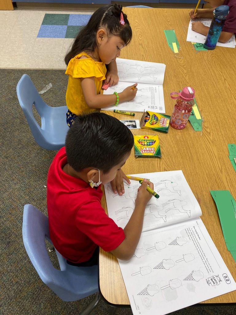 Two kindergarten students sitting at their desk tracing in crayon a shape in their workbook.